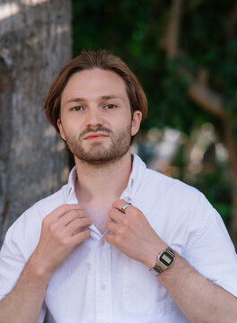Young handsome man in a white short sleeve shirt adjusting his collar, outdoors male portrait with green background.