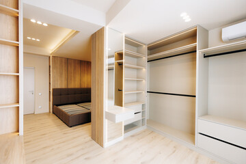 Fototapeta premium Bright modern walk-in closet interior with open shelving drawers and hanging rails, integrated vanity with mirror, connected bedroom view, neutral tones and recessed ceiling lights