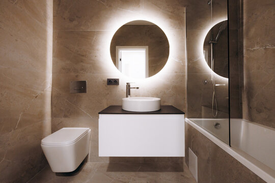 Contemporary beige marble bathroom interior with warm backlit illuminated round mirror, floating vanity vessel sink, chrome fixtures, and bathtub with glass shower screen