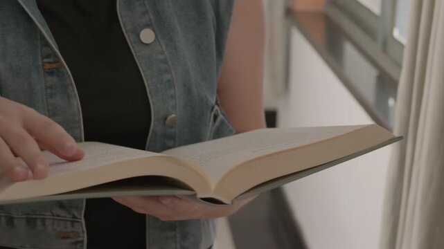 A person holds an open book while reading indoors, wearing casual clothing near a window.