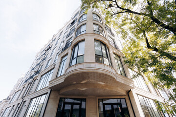 Limestone corner building with curved bay windows and glass panes under green tree canopy in bright daylight, low-angle city view of elegant exterior facade © Kantemir Production