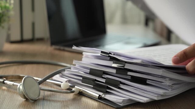 Stethoscope beside stacked paperwork with person working on laptop, showing medical records management and healthcare documentation workflow in an office.