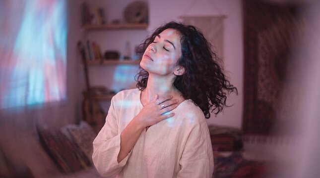Woman with hand on chest and eyes closed in soft colorful light, breathing, mindfulness, self-care, gratitude, emotional healing, cozy bedroom