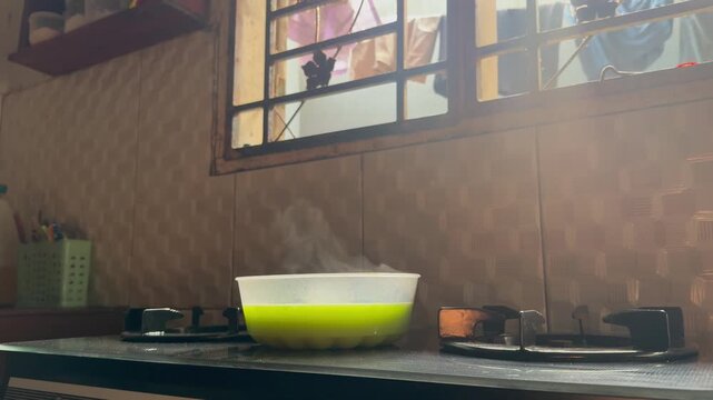 Green liquid bubbles in a kitchen appliance. Bubbles rise in a clear bowl on a stovetop. The appliance sits atop a modern cooker. Kitchen tiles have a subtle geometric pattern