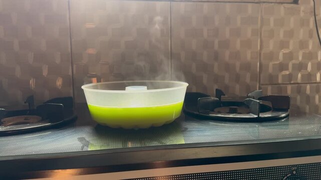 Green liquid bubbles in a kitchen appliance. Bubbles rise in a clear bowl on a stovetop. The appliance sits atop a modern cooker. Kitchen tiles have a subtle geometric pattern