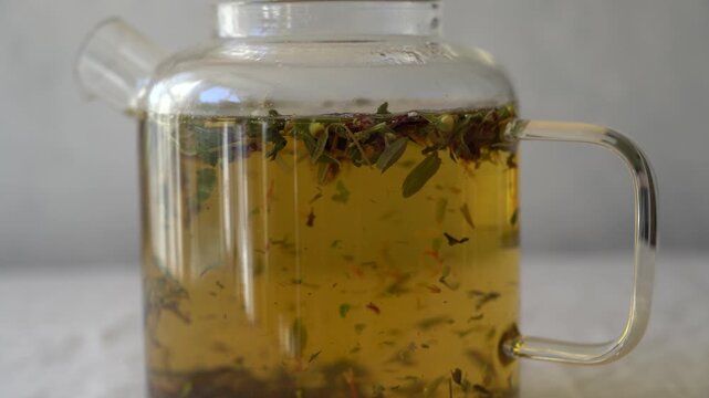 Slow motion herbal tea brewing in a transparent glass teapot, dried herbs infusing in hot water, natural beverage close up
