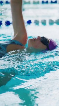 Swimmer glides on backstroke through clear pool water. Purple cap and goggles highlight focused effort. Lane markers guide steady movement. Ideal for fitness, sports, or wellness projects