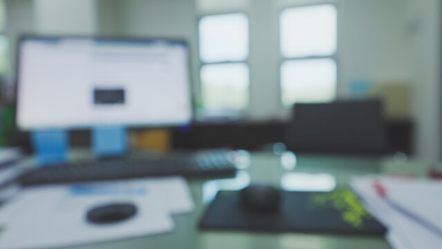 Refocused view shows corporate desk setup featuring computer monitor, keyboard, paperwork, mouse pad, chair, windows, creating empty workspace setting suited as background.