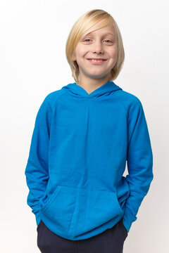 Young blond boy standing, smiling happily at the camera, wearing a casual blue hooded sweatshirt with his hands in the front pocket against a white background