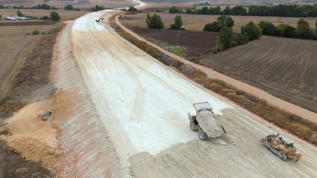 An aerial view of heavy machinery including a dump truck unloading soil, a bulldozer, and a grader working on a road construction embankment