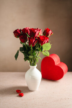 Valentine's Day festive background. Red roses flowers in a vase and heart gift box on warm background. Festive greeting card, flat lay
