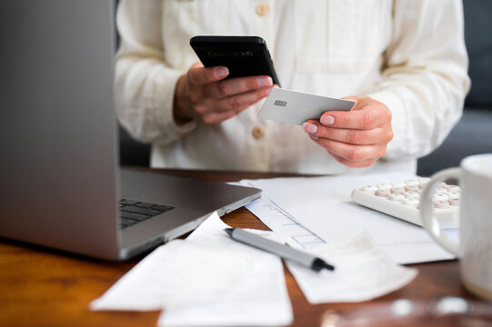 Woman using a smartphone and bank card for an online purchase at a laptop, representing ecommerce, digital payments, mobile shopping, consumer transactions and convenient cashless spending.