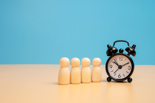 Four wooden human figures standing in a row beside a black alarm clock on a beige surface against a blue background. Concept of waiting time, team scheduling, deadlines and workforce time management.