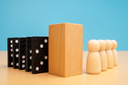 Upright wooden block separating black domino tiles from a group of wooden human figures. risk prevention, protective barriers and shielding people against a blue background.