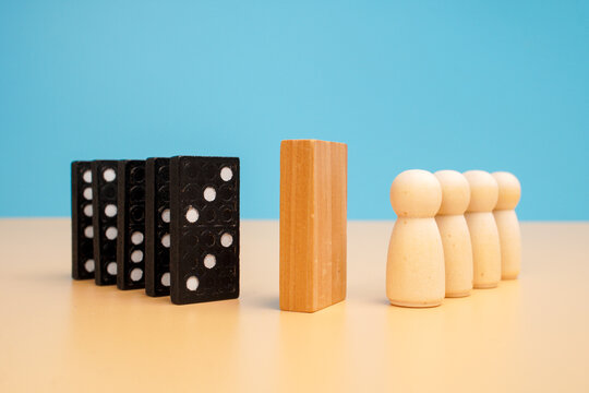 Black domino tiles positioned opposite wooden human figures with an upright wooden barrier between them. Protecting communities, interrupting chain reactions and managing collective risk.