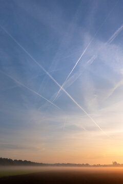 Lots of contrails in an orange blue sky at sunrise above fields ground fog