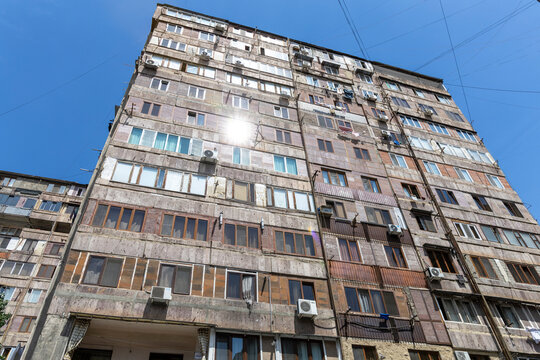 Low angle view of a soviet era apartment tower with sun flare shining between rows of windows and tangled electric wires against a bright blue sky, Yerevan, Armenia