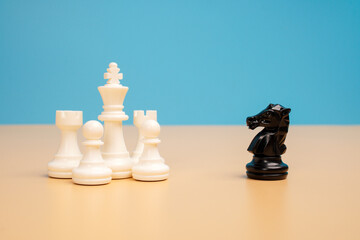 A black chess knight faces a group of white chess pieces on a beige surface against a blue background. Concept of competitive challenges, strategic thinking, courage and facing stronger opposition. © Akmalism Photography