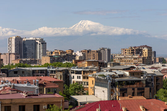 City skyline of red roofed apartment buildings with the snow capped peak of Mount Ararat rising above the clouds on the horizon in soft morning light, Yerevan, Armenia