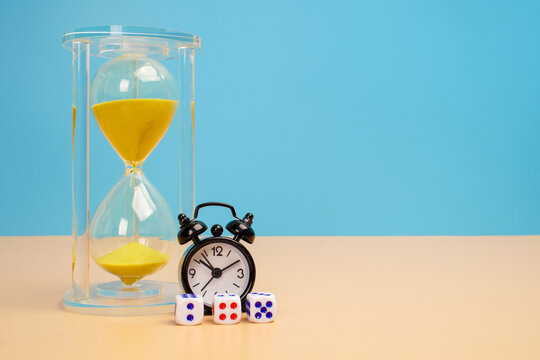 Yellow sand hourglass with a small black alarm clock and dice arranged at its base on a beige surface against a blue background. Countdown, deadline pressure, uncertainty and timely decision making.