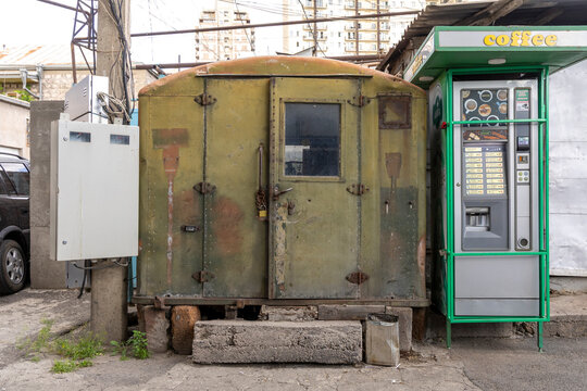Old rusty metal kiosk locked with a chain and padlock standing beside a green coffee vending machine on a weathered city street near parked cars, Yerevan, Armenia