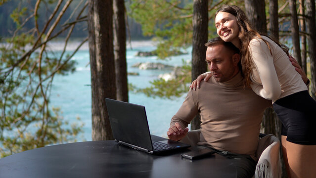 Beautiful romantic young couple kissing and hugging during remote work on an outdoor hotel terrace near a turquoise river.