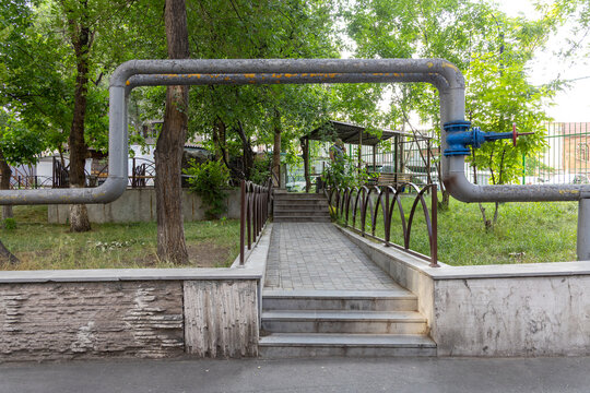 Overground heating system pipes forming a rusted metal arch above a paved walkway lined with trees inside a quiet green residential courtyard, Yerevan, Armenia