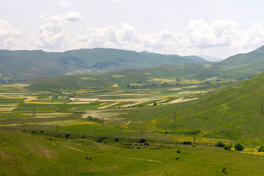 Aerial view of a green valley with patchwork farmland rolling hills and a railway track winding through the tranquil countryside beneath distant mountains and sky, Haghpat, Armenia