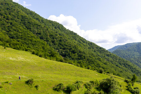 A lone hiker carrying a backpack walks across a green mountain meadow surrounded by forested hills beneath a wide open sky on a scenic trekking adventure, Haghpat, Armenia