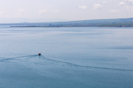 Panoramic view of Lake Sevan with a small boat sailing calmly across tranquil blue waters beneath distant snow capped mountains and a vast highland landscape, Sevanavank, Armenia
