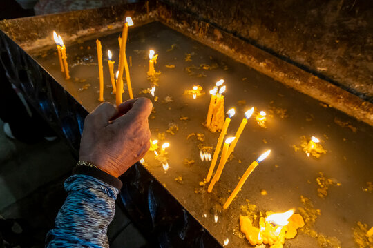 Close up of a hand lighting a single candle among many burning candles glowing warmly with melted wax inside the candlelit interior of Sevanavank Monastery, Sevanavank, Armenia