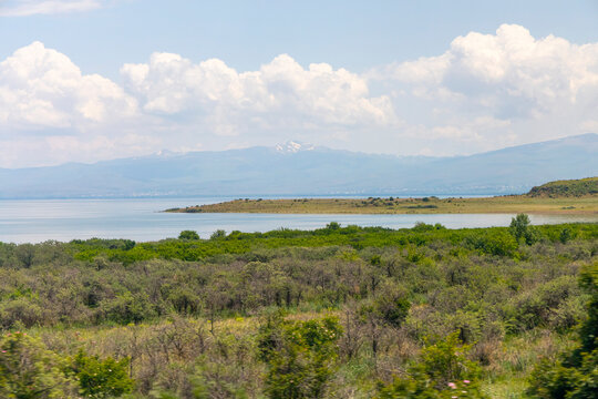 Snow capped mountains rise above a peninsula of green shrubland and wild vegetation along the shore of the lake under a scenic highland sky in Gegharkunik Province, Sevan, Armenia