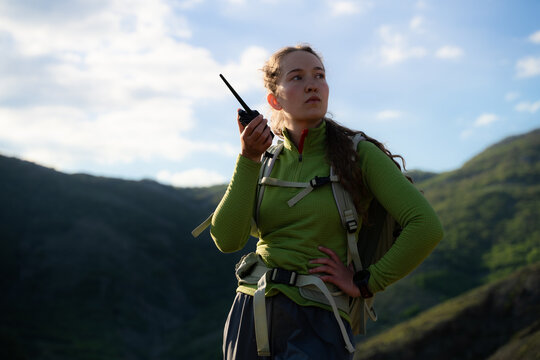 Young woman in hiking clothing using a walkie-talkie during an outdoor trek, surrounded by a dramatic mountainous landscape and open sky.