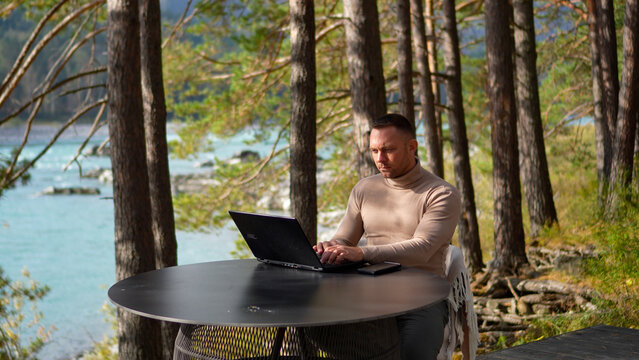 Side view of a concentrated male freelancer typing on a laptop during remote work at a scenic wooden deck terrace in a pine forest.
