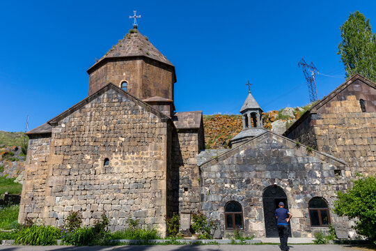 Man wearing a cap walks along a stone pathway toward the arched entrance of Vanevan Monastery past ancient stone buildings and a carved khachkar, Artsvanist, Armenia