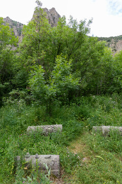 Ancient Jewish gravestones lie scattered across the grass below a rocky cliff surrounded by forest trees and wildflowers in a quiet mountain wilderness, Eghegis, Armenia