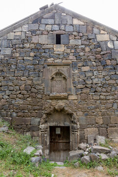 Carved khachkar cross stone is set above the arched entrance of an ancient stone church beside a wooden door and weathered gable facade in the countryside, Eghegis, Armenia