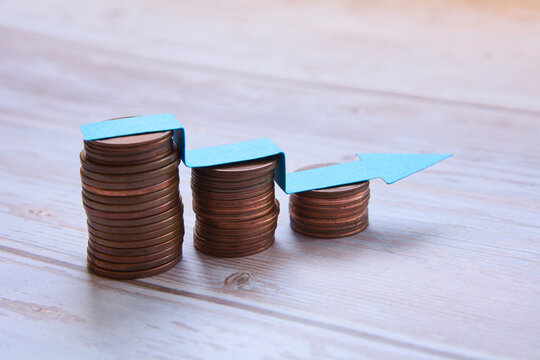 Decreasing stacks of copper coins on a light wooden table with a blue paper arrow descending step by step, concept of economic decline and shrinking savings