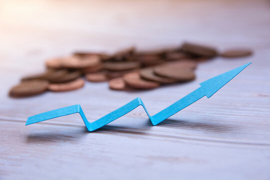 Blue paper arrow zigzagging upward in front of a blurred pile of copper coins on a wooden table, concept of financial growth, recovery and rising profits