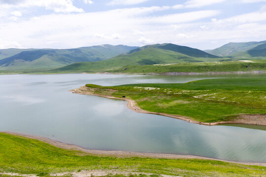 Scenic mountain reservoir reflects a calm blue sky while surrounded by green rolling hills and a lone car parked along its quiet remote shoreline, Tatev, Armenia