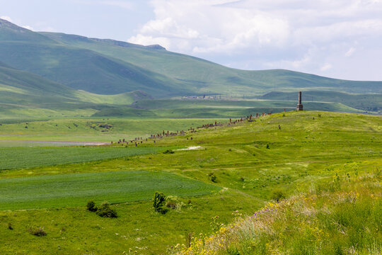 Old cemetery with rows of carved khachkar stone monuments and a tall obelisk stands peacefully in a green valley surrounded by distant rolling hills, Tatev, Armenia