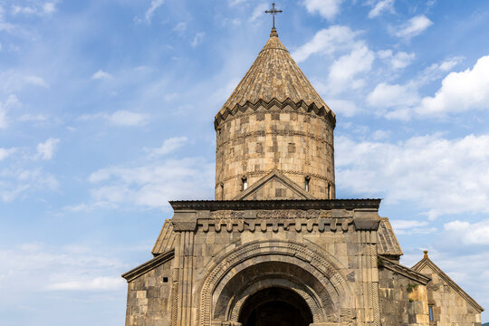 The facade and cylindrical bell tower of Tatev Monastery feature a carved stone entrance arch with ornate patterns set beneath a bright clear blue sky, Tatev, Armenia