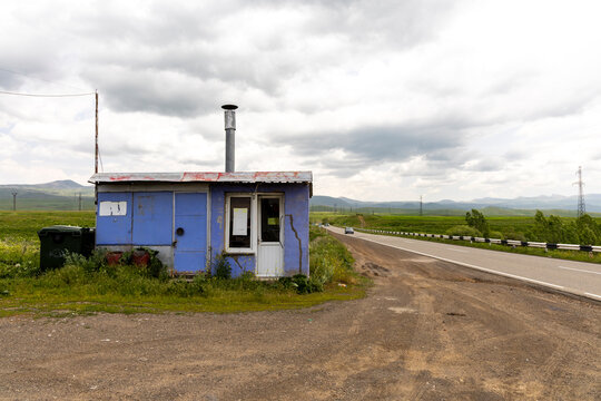 Small weathered blue roadside shop with a rusty chimney and faded signboard standing empty along a quiet countryside highway beneath rolling hills, Noravank, Armenia