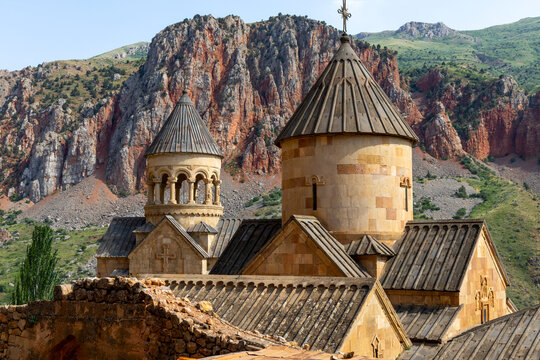 Conical domes and a stone belfry of the medieval Surb Astvatsatsin church rise with arches and carved columns against a backdrop of dramatic red canyon cliffs, Noravank, Armenia