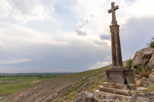 A carved stone cross monument stands on a hillside overlooking the snow capped peak of Mount Ararat beyond vineyards wheat fields and a wide valley plain, Khor Virap, Armenia