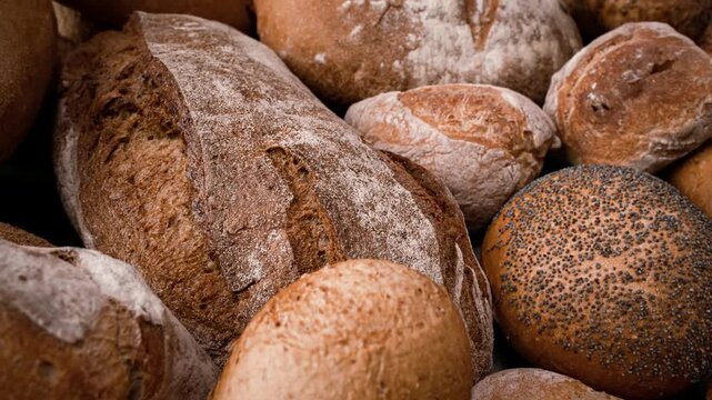 Camera glides over assortment of artisan breads featuring sourdough loaves and poppy seed buns in warm lighting.