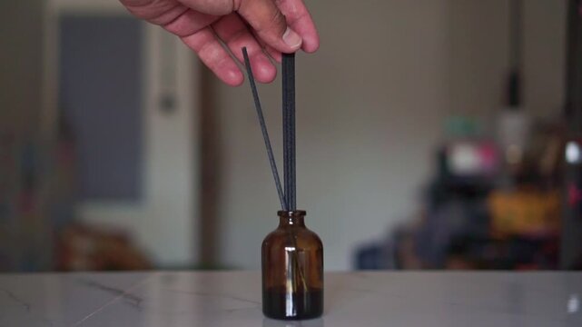 Hand arranging reed sticks in an amber glass diffuser bottle on a marble table. Home fragrance footage showing aromatherapy, relaxation, and interior decor.