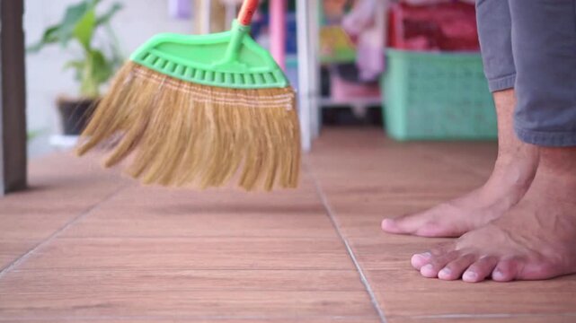 Barefoot person sweeping a wooden tile floor with a green broom indoors. Household footage showing cleaning, housework, and daily domestic routine.