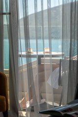 Fototapeta premium Woman in a white bathrobe relaxing on a private hotel balcony overlooking the blue sea and mountains. View through sheer curtains creates a peaceful morning vacation and wellness atmosphere.