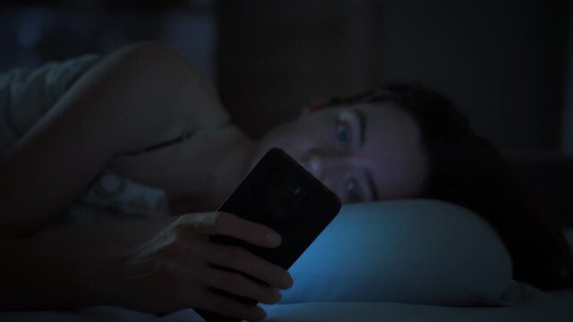 A young woman lies in bed in a dark bedroom facing the camera. Smartphone screen lights up her face in the dark. Concept of insomnia caused by social media and late night phone use